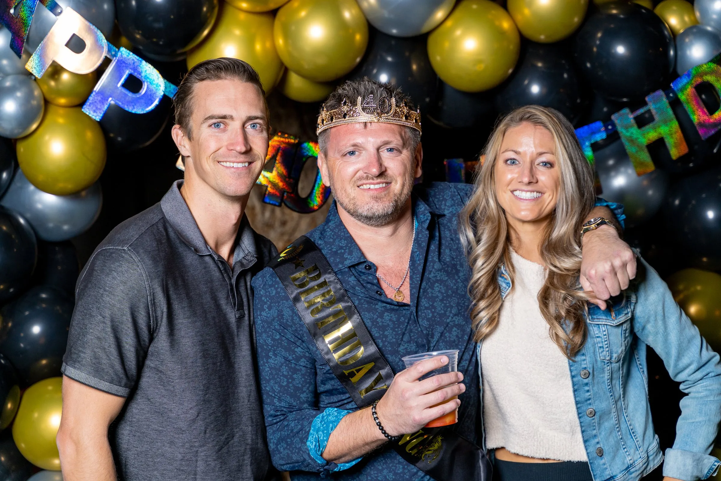 Three people, one in a crown and sash, pose before a black and gold balloon garland.