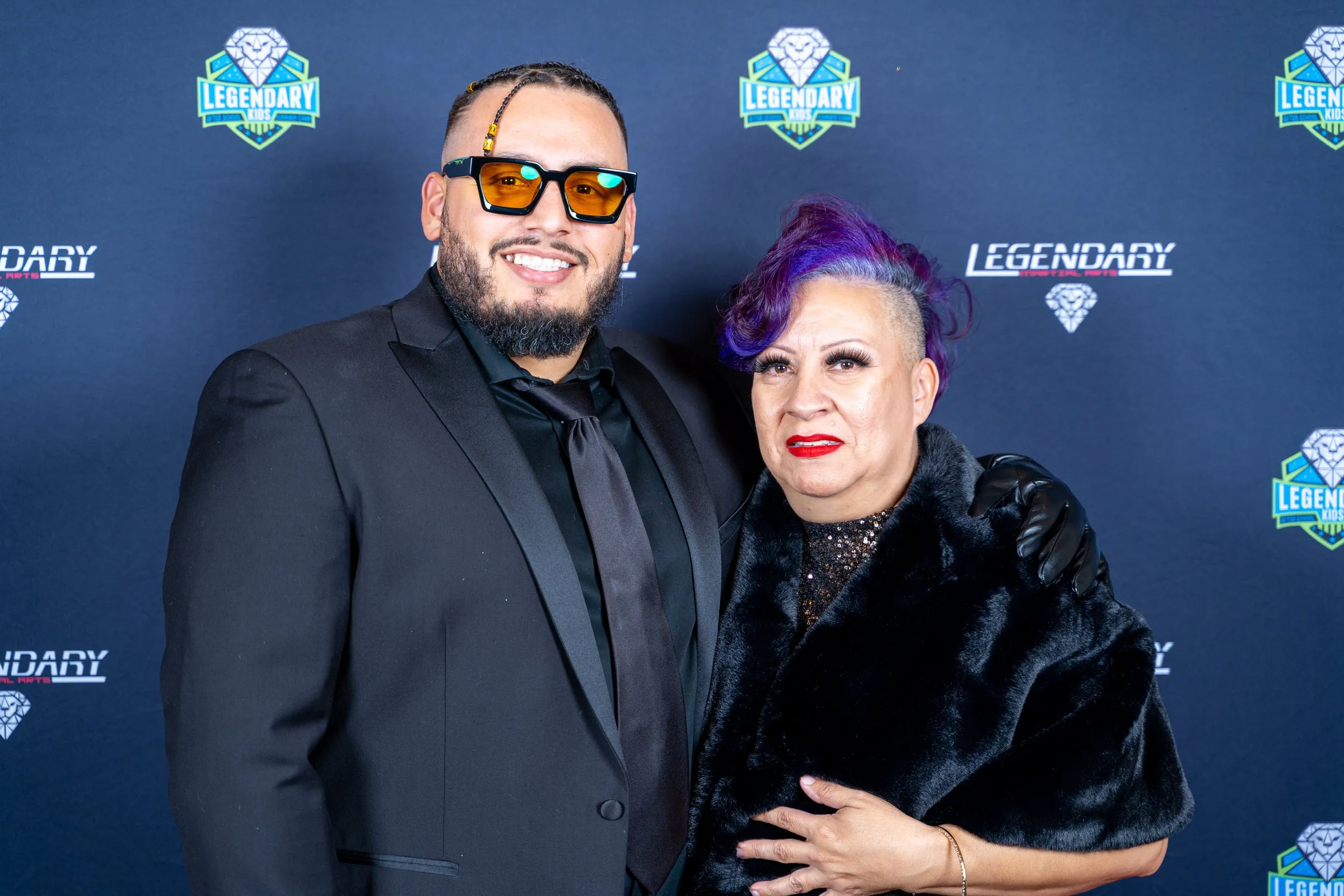 Man in orange-tinted sunglasses and woman with purple hair pose close before a navy backdrop.