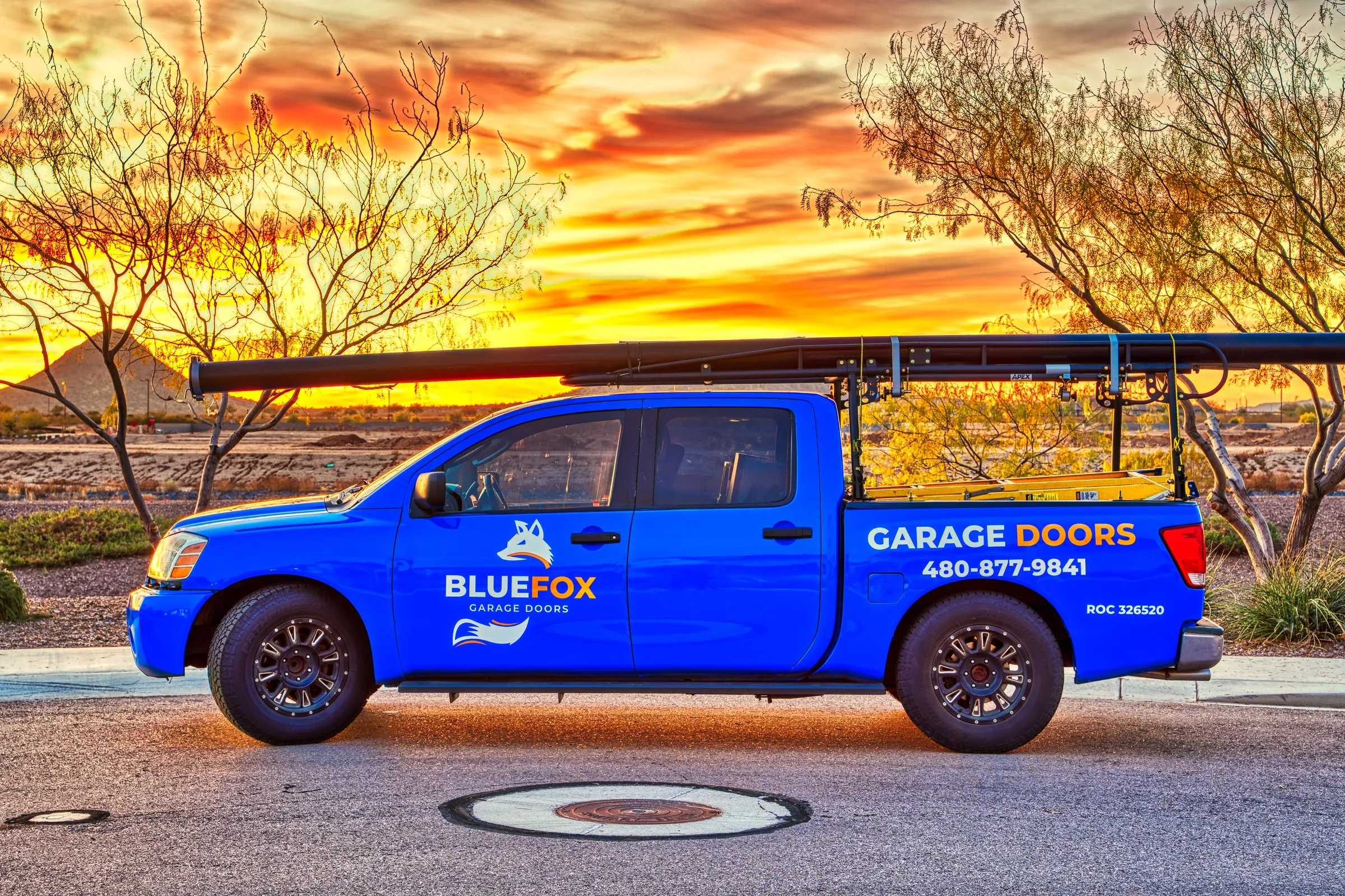 Blue work truck with a ladder rack parked beneath a fiery orange sunset sky.