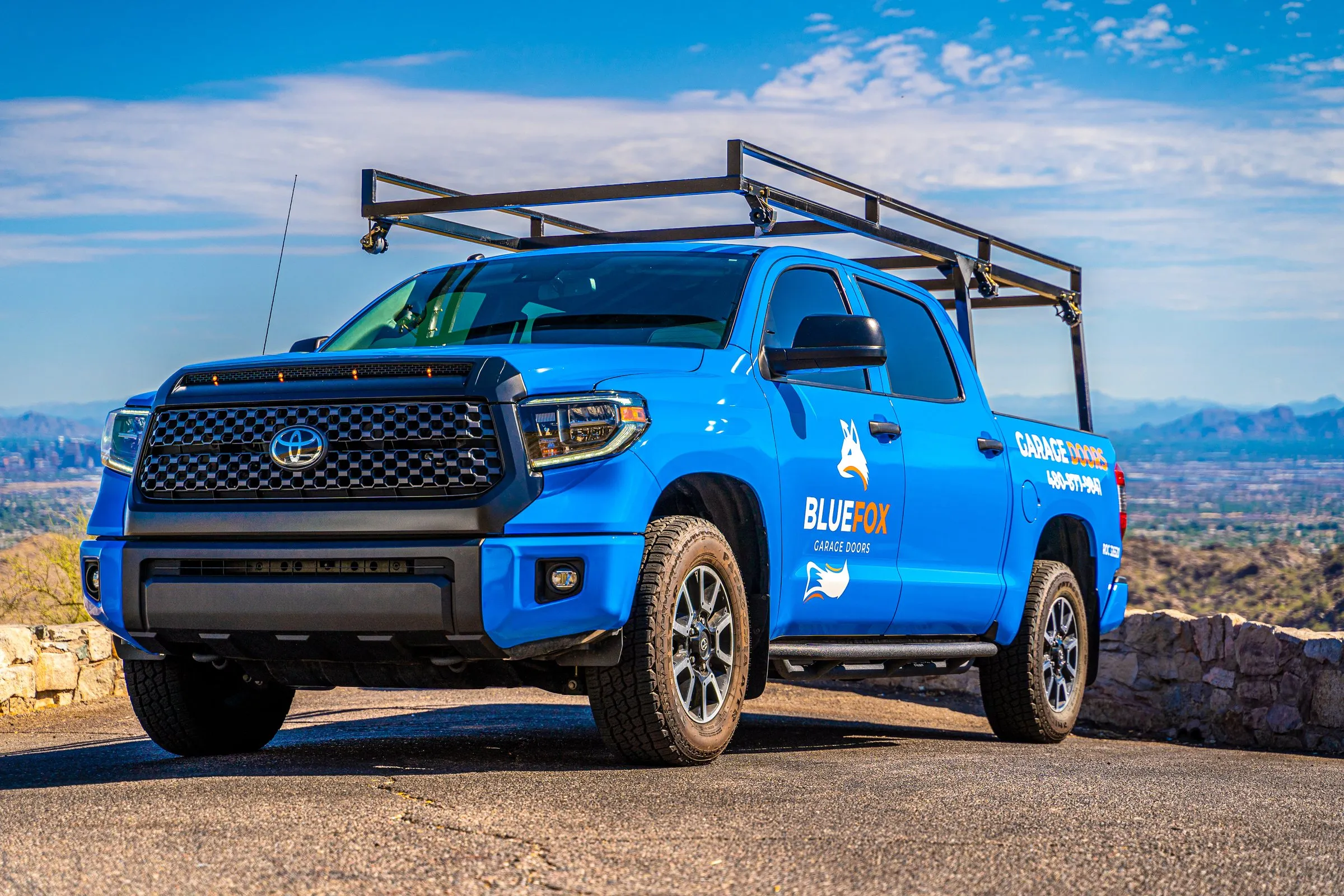 Blue pickup truck with a roof rack parked at a scenic overlook under a bright sky.