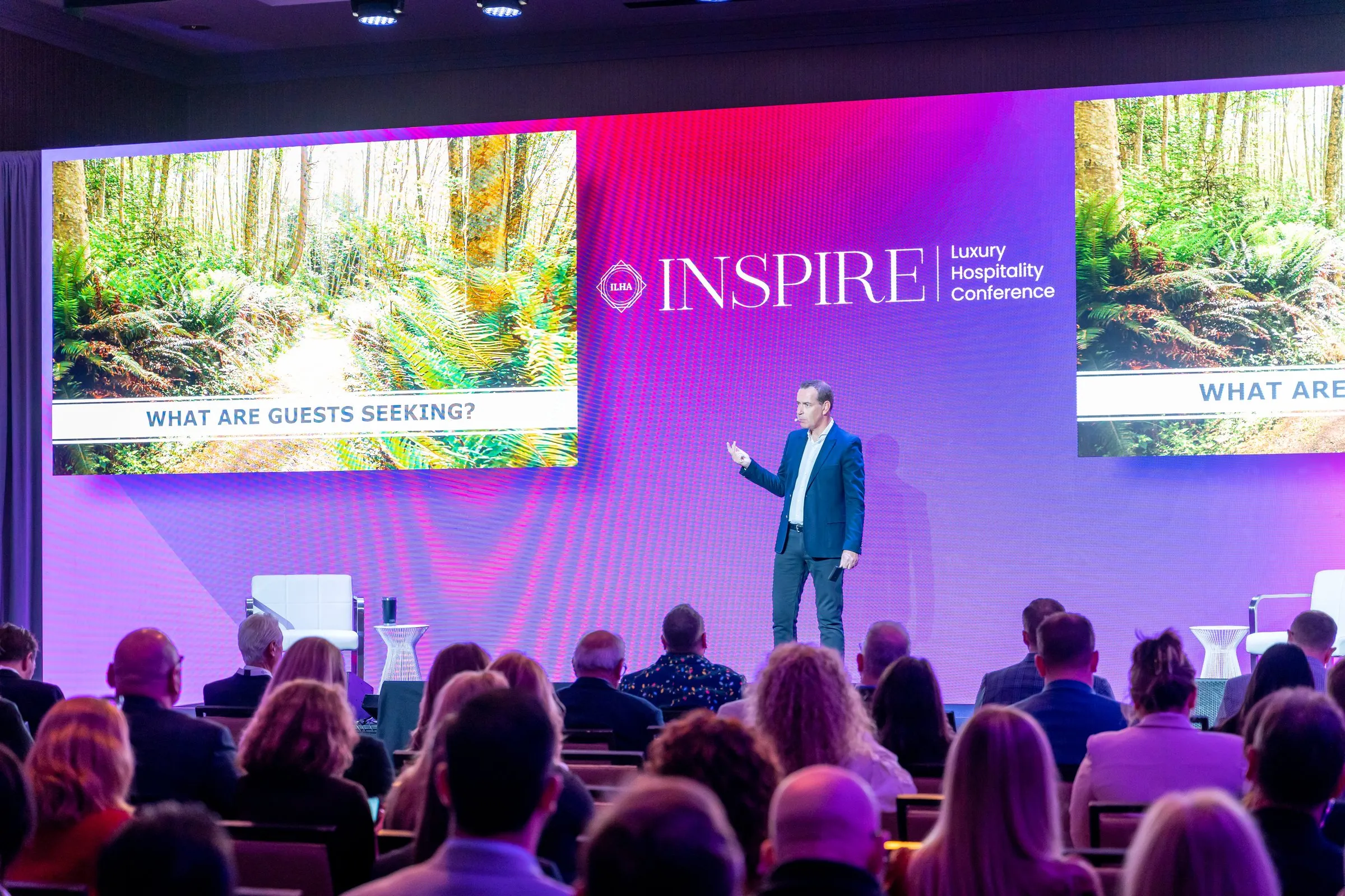 Speaker stands on a magenta-lit conference stage before a seated audience and large screens.