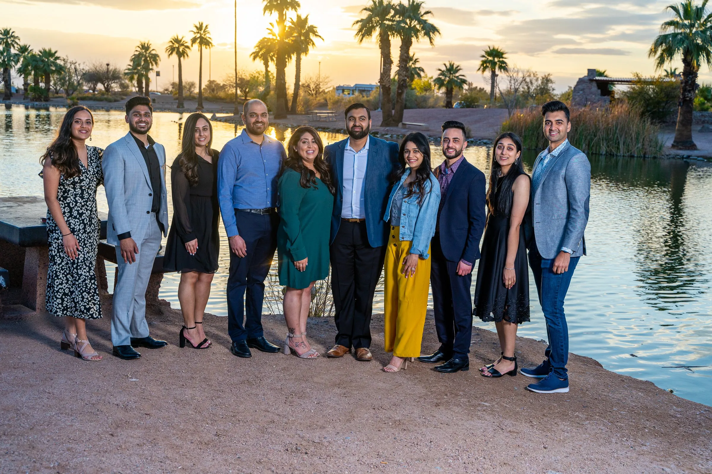 A group of about a dozen people in business-casual outfits pose beside a lake at sunset.