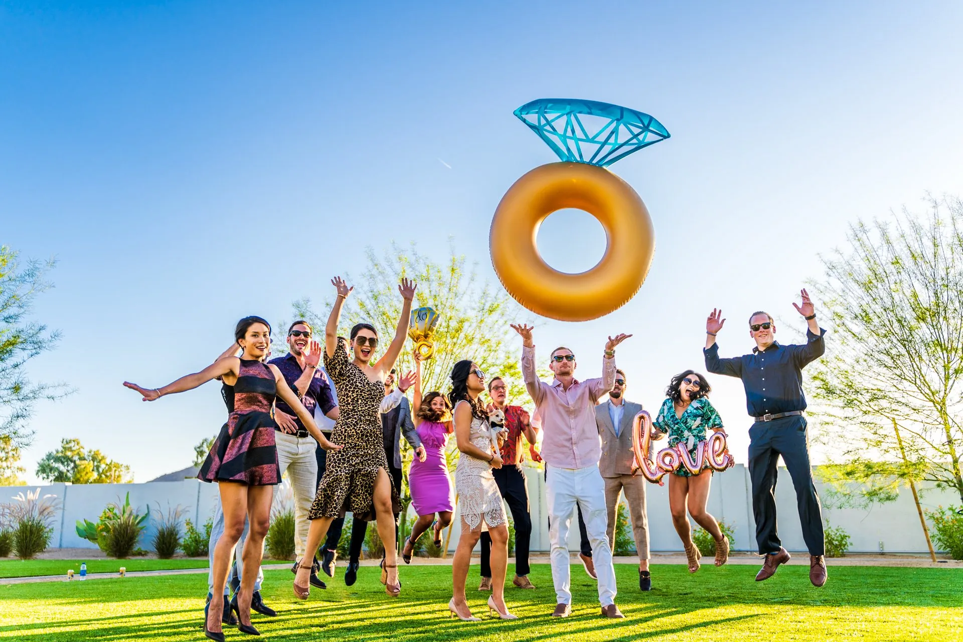 Group of people leap beneath a giant inflatable diamond ring on a sunny lawn.