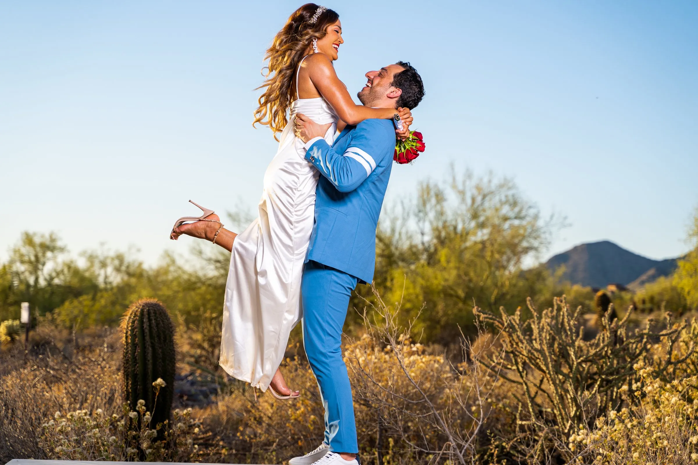 Groom in a sky-blue suit lifts his laughing bride, heel kicked up, among desert cactus.
