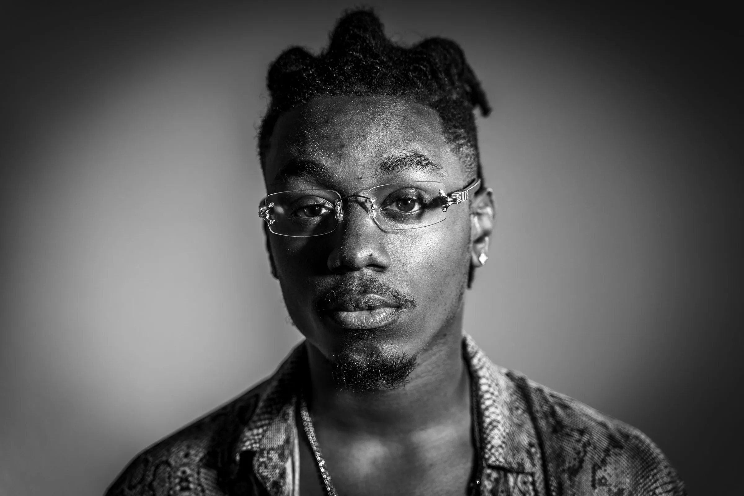 Black-and-white portrait of a young man with glasses and styled hair, dark vignette edges.