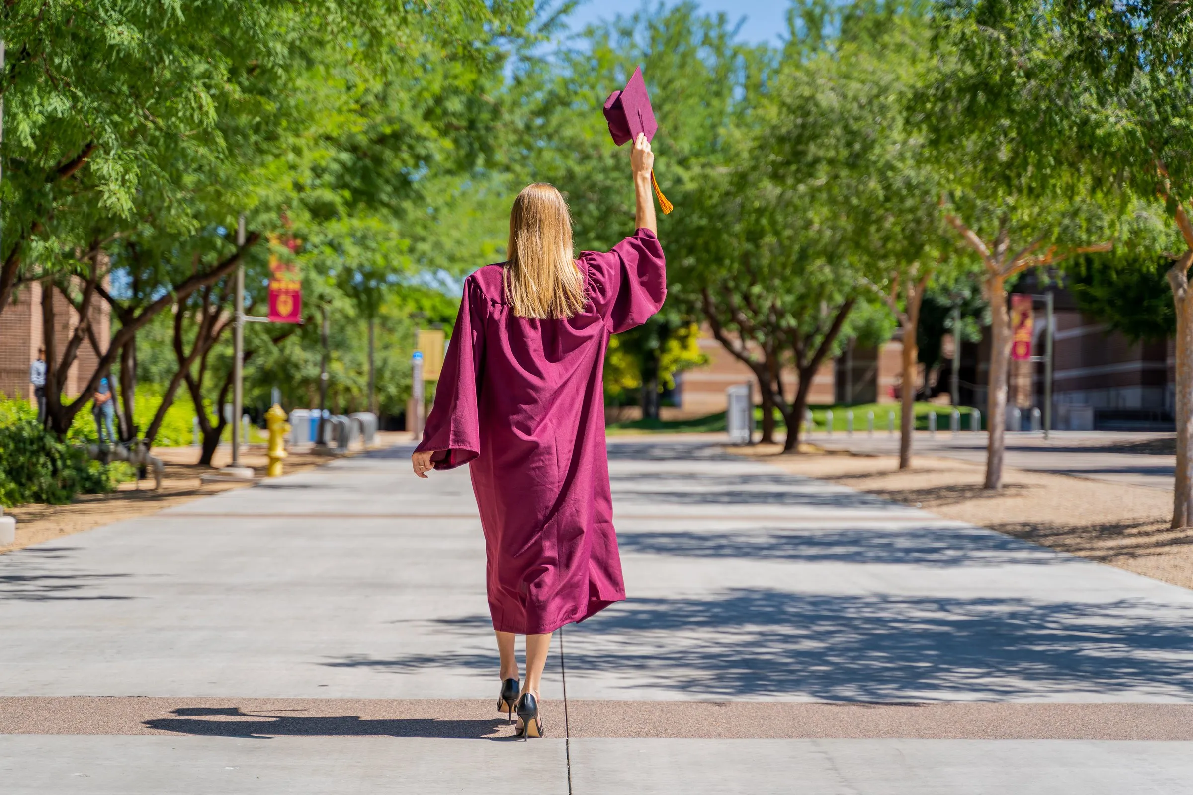 Graduate in a maroon gown walks away along a tree-lined campus path, cap raised.