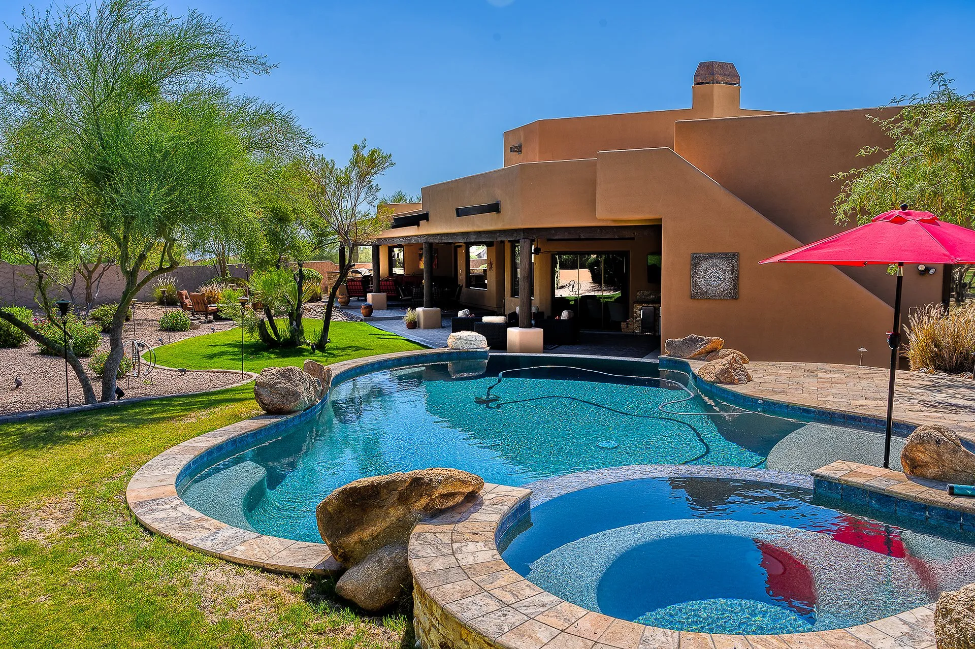 Southwestern-style home with a curving backyard pool, spa and red patio umbrella under blue sky.